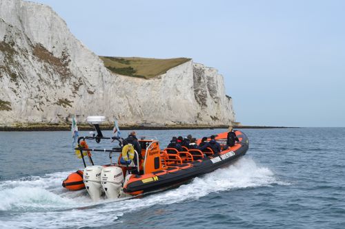 People aboard a black and orange Dover Sea Safari RHIB heading past the White Cliffs on a blue sea with blue sky overhead.