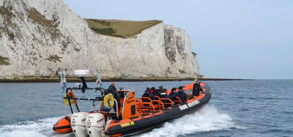People aboard a black and orange Dover Sea Safari RHIB heading past the White Cliffs on a blue sea with blue sky overhead.