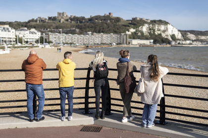 A group of five people leaning on railings looking over Dover beach towards Dover Castle.