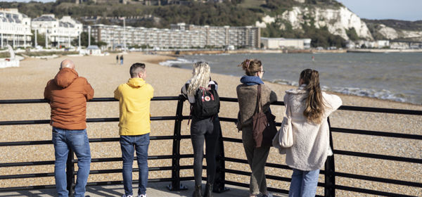 A group of five people leaning on railings looking over Dover beach towards Dover Castle.