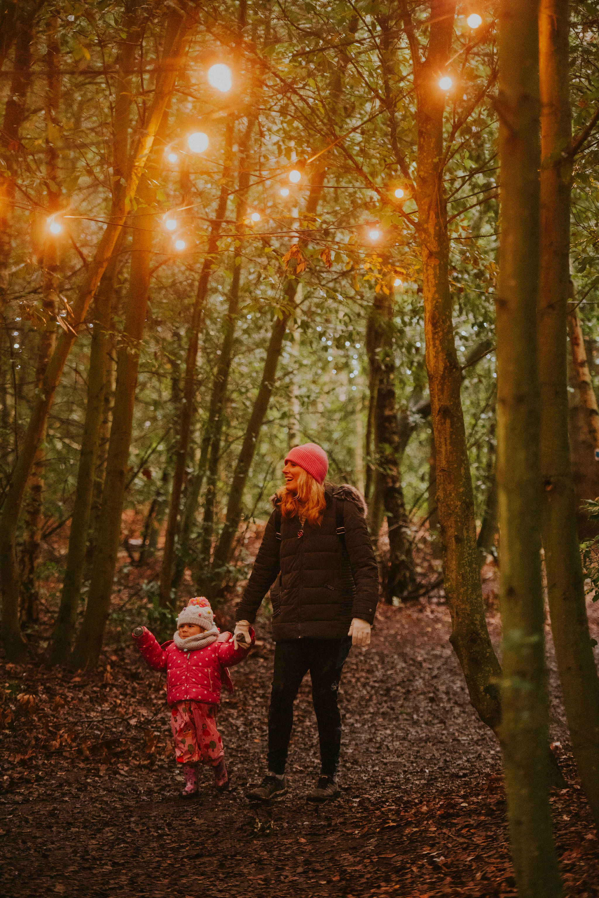 A woman and small child in winter coats walking through woods with lights