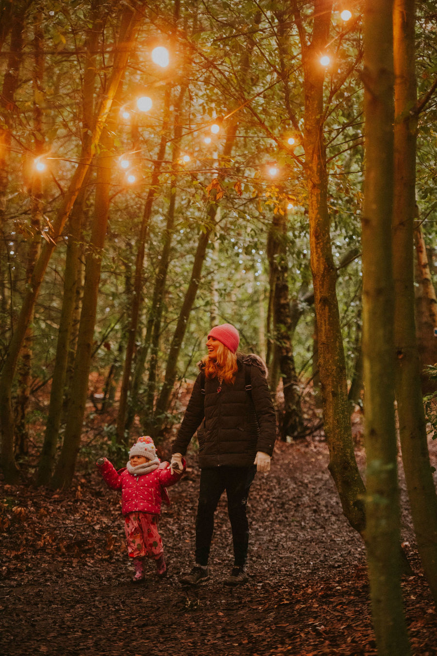 A woman and small child in winter coats walking through woods with lights