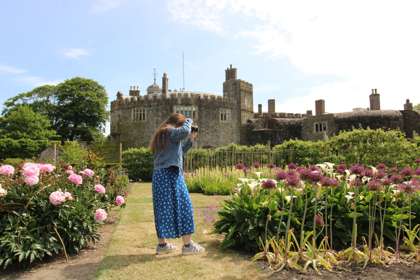 A woman in a blue dress photographing flowers with Walmer Castle in the background.