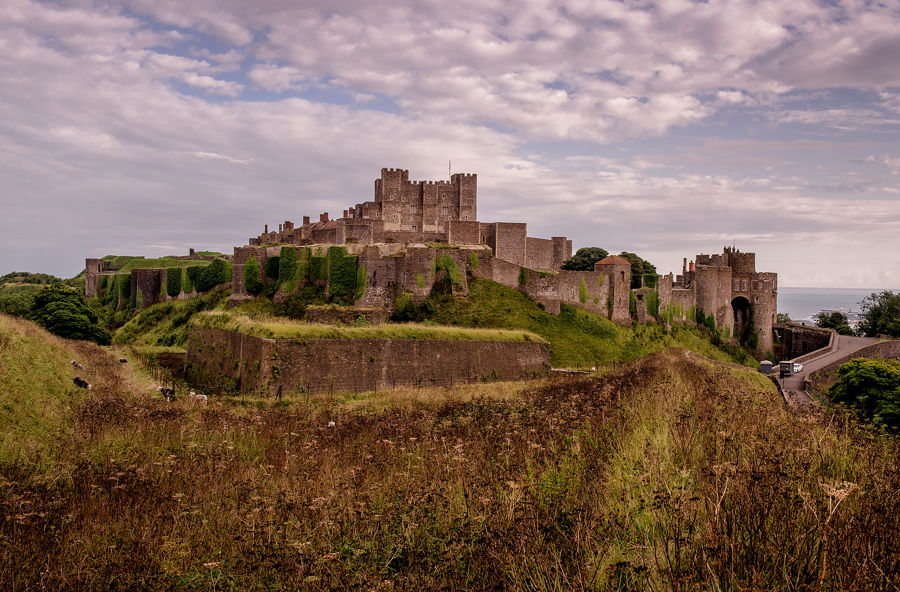Dover Castle from a distance with greenery in the foreground, cloudy sky and sea just visible
