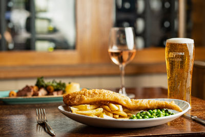 A plate of fish and chips, a pint of beer and a glass of wine on a table. 