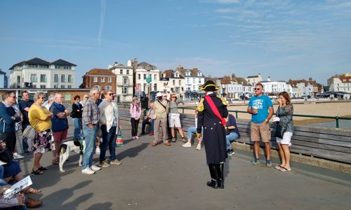 A man dressed in Napoleonic costume talking to a group of tourists on Deal Pier with the beach and seafront in the background.