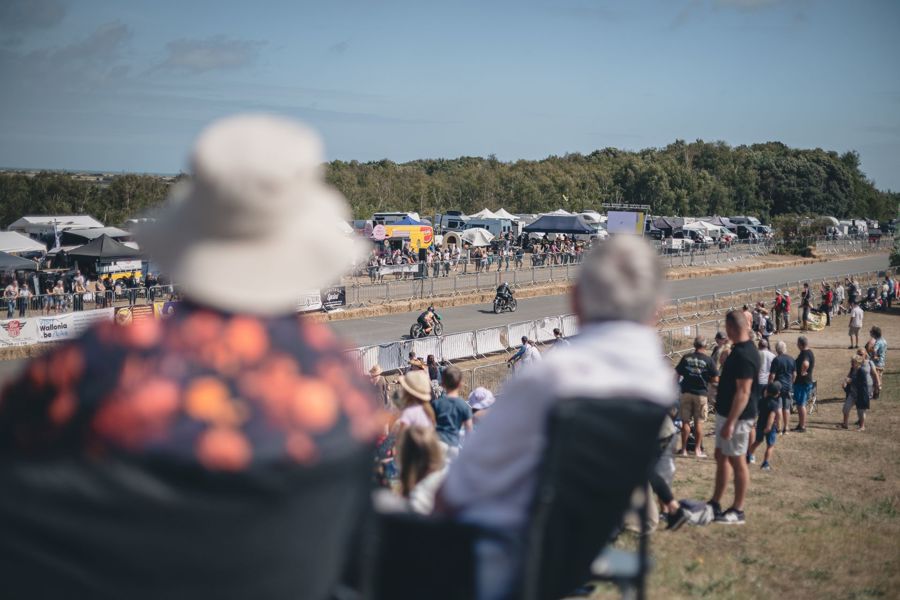 A crowd of people watching vintage motorbike racing with stalls in the background