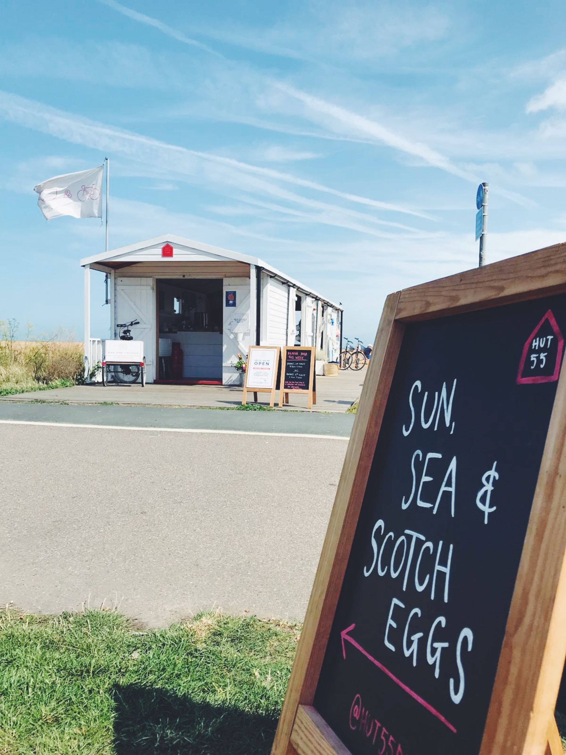 A chalkboard sign saying 'Sun, Sea & Scotch Eggs' pointing at a beach hut cafe, Hut 55.