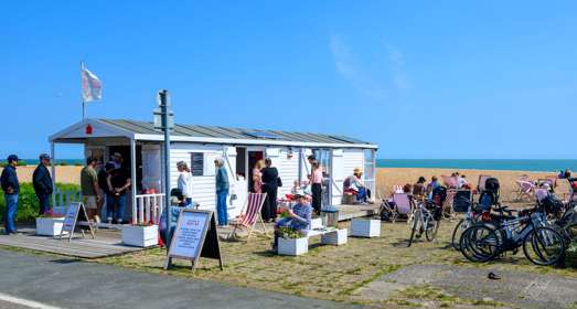 A beach hut cafe with people sitting on red and white striped deckchairs and rows of bikes
