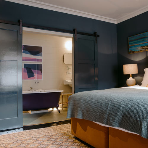 Interior of a hotel bedroom with dark walls and a mustard-coloured bedstead, with sliding doors revealing an ensuite bathroom with roll-top bath.