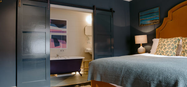 A bedroom with mustard coloured bed, dark grey walls and open door revealing a roll-top bath in the en-suite bathroom. 