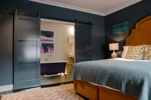 Interior of a hotel bedroom with dark walls and a mustard-coloured bedstead, with sliding doors revealing an ensuite bathroom with roll-top bath.