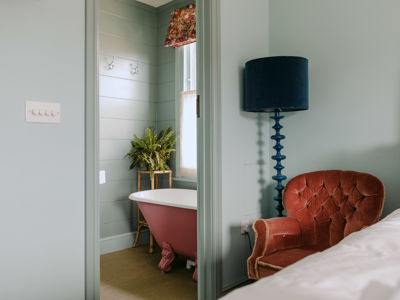 A pale blue bedroom with pink armchair, dark blue standard lamp and doorway to bathroom with pink roll-top bath.