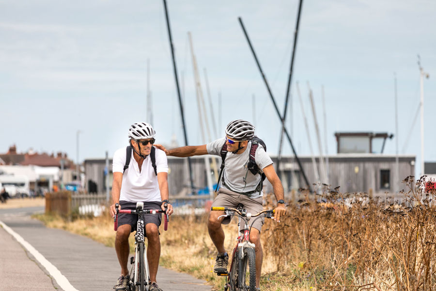 Two men cycling, one with his arm on the other, along the coast path with Downs Sailing Club in the background