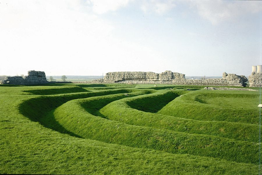 Green grass ditches with the remains of Roman walls in the background at Richborough Roman Fort.