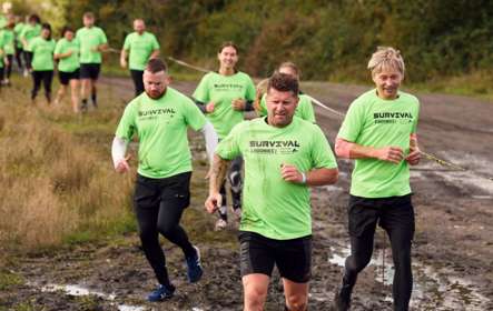 A group of people wearing green Survival t-shirts run through the mud along the course