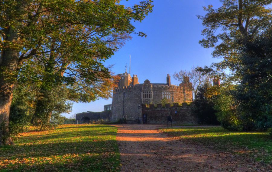 A path, covered in fallen leaves bordered by trees, to Walmer Castle with a blue sky overhead.