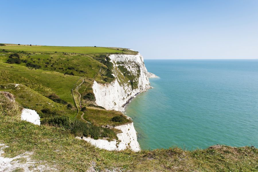 A clifftop view along the White Cliffs of Dover on a sunny day showing the coast path, chalk cliff face and blue sea to the right.