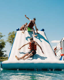 Young people wearing red life jackets and swimsuits sliding down a giant, floating, inflatable slide into blue water.