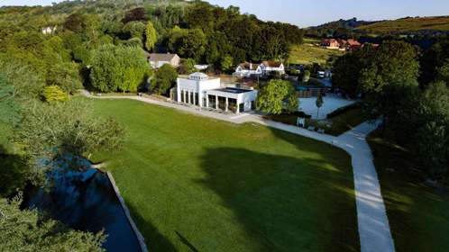 the white buldings of Kearsney Cafe nestled into the green park of Kearsney Abbey with mature trees in leaf, green grass, and rolling hills in the distance. 