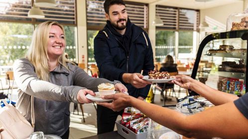 Two people at a cafe counter being handed cakes by the server.