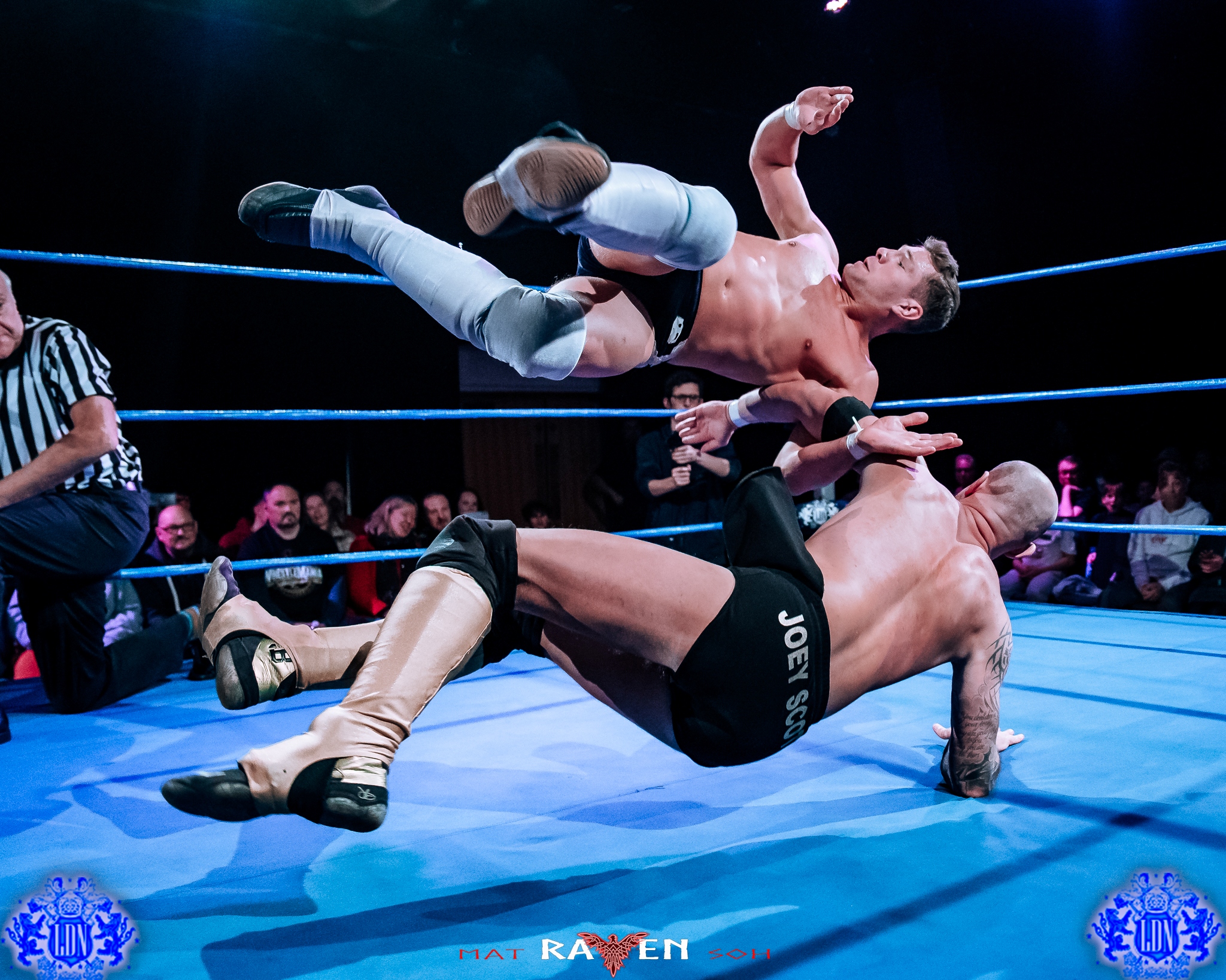 2 men wrestling in a wresting ring with a referee down on one knee
