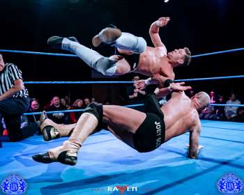 2 men wrestling in a wrestling ring watched by a referee 