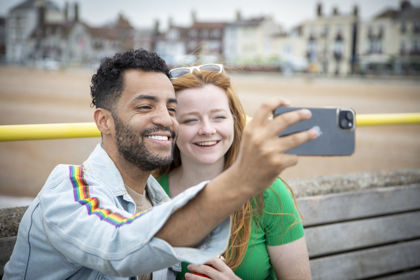 Two people taking a selfie with a smartphone with Deal beach and seafront in the background.