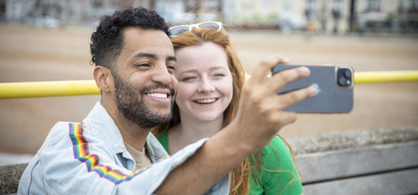 Two people taking a selfie with a smartphone with Deal beach and seafront in the background.
