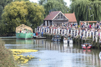 The River Stour and Sandwich Quay with crowds of people lining the riverbank to watch the annual duck race (yellow rubber ducks and boats in the water). Sandwich Medieval Centre in the background. 