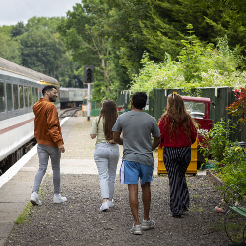 Four people on a station platform with a train to the left of the image and a green fence and trees to the right.