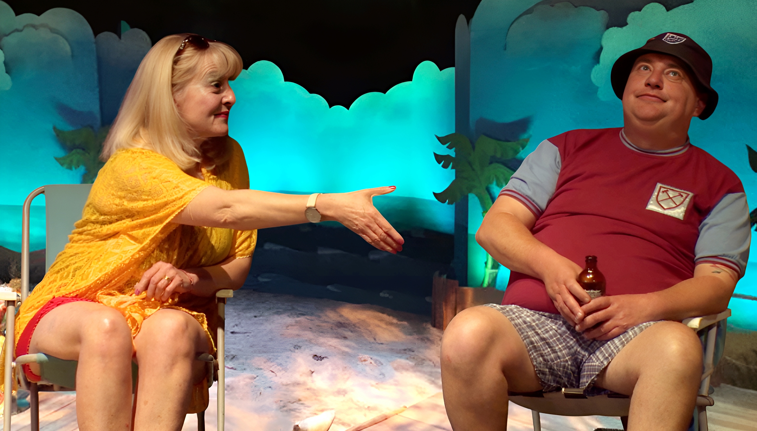 2 actors sitting on deck chairs as though on a Greek island the man is holding a bottle of beer