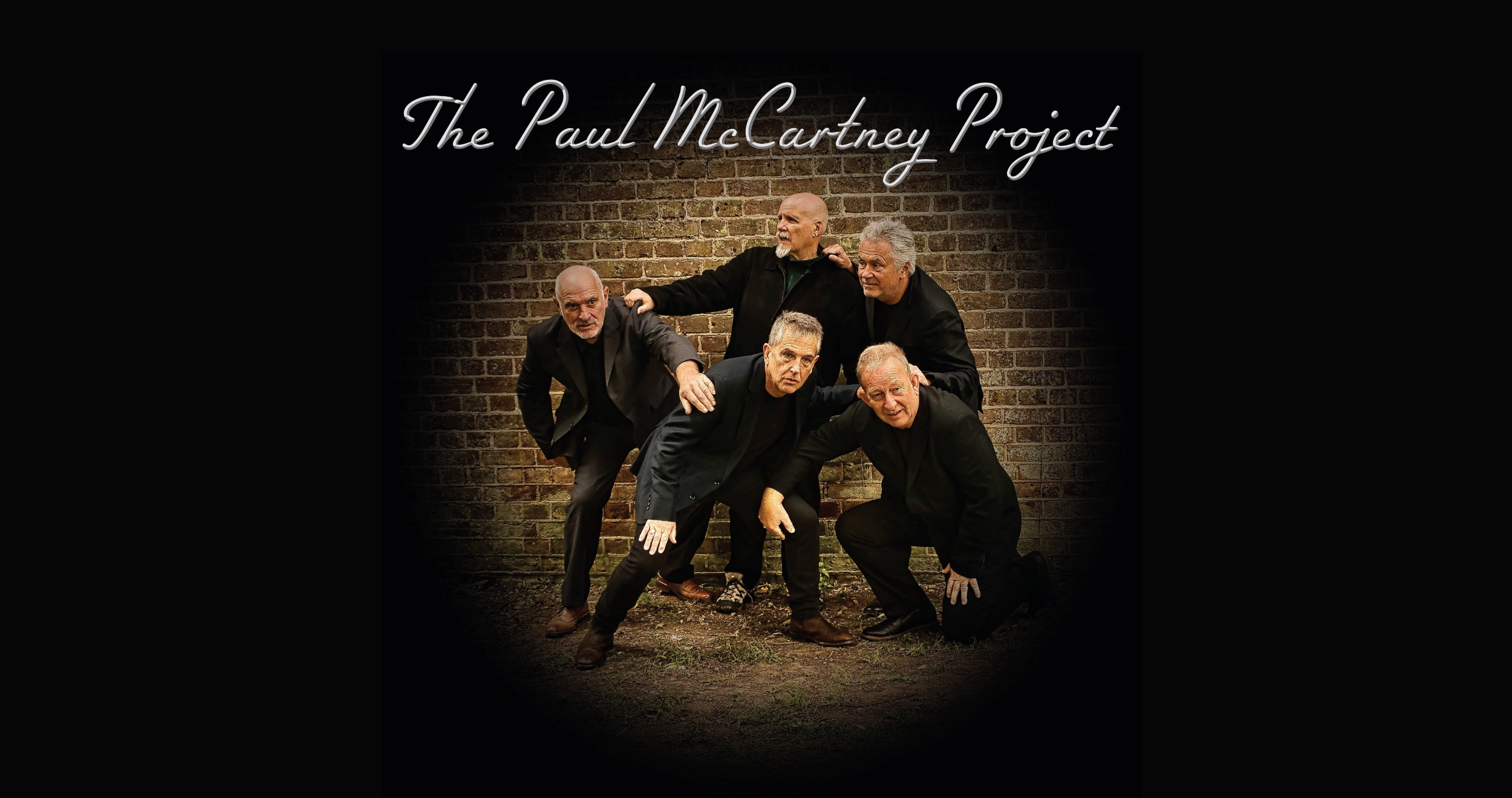 The 5 members of the band re-create the Band on the run album cover crouching in front of a brick wall
