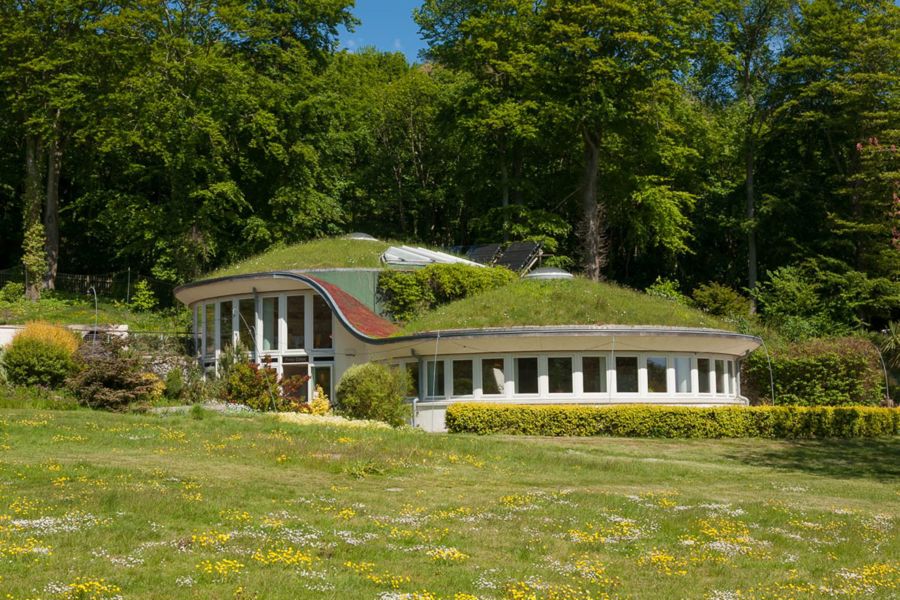 A meadow in the foreground with a circular building with green roof and green trees behind.