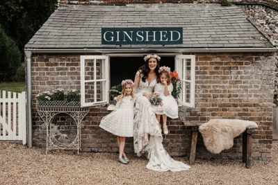 A smiling bride with two small bridesmaids sitting in a window of a brick gin shed.