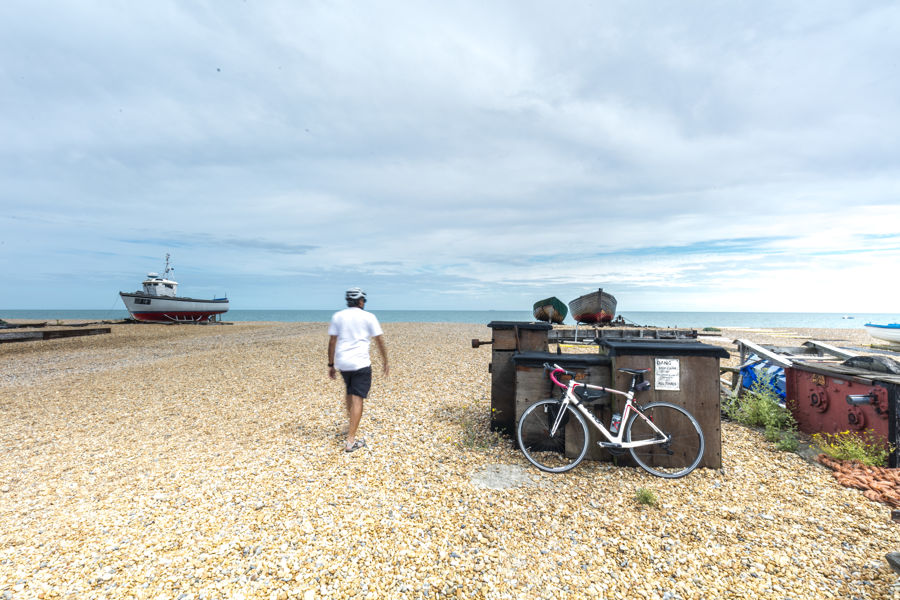 A bicycle propped against a wooden storage box on a shingle beach, with the rider walking towards the sea in the distance and a few boats.