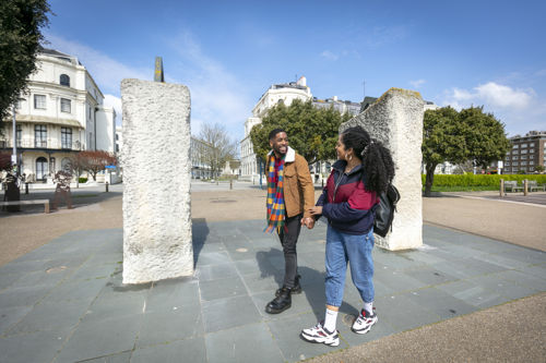 A couple walking past the sea-swimmers sculpture on Dover promenade with Victorian buildings in the background.