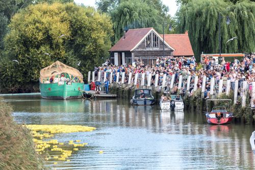 A crowd of people on a riverbank with boats moored along the quayside and a red roofed building.
