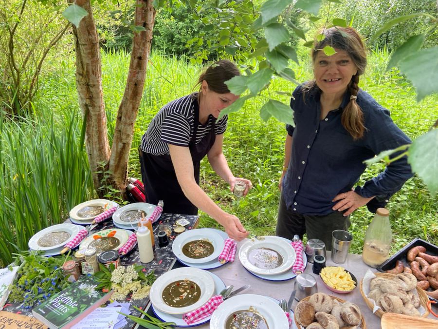 Two women behind a table laid with foraged foods in a woodland environment.