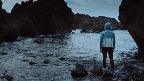 A person wearing blue hooded coat and black trousers looking out over rocks and sea