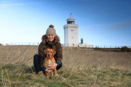 A woman wearing a coat and bobble hat crouched with a brown dog with a red collar both looking at the camera. The South Foreland Lighthouse and a blue sky behind.