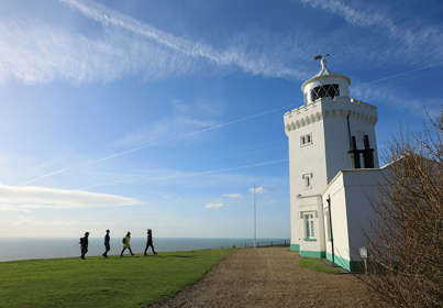 4 people approaching a white painted lighthouse on foot, across a green lawn with the sea just visible in the background.