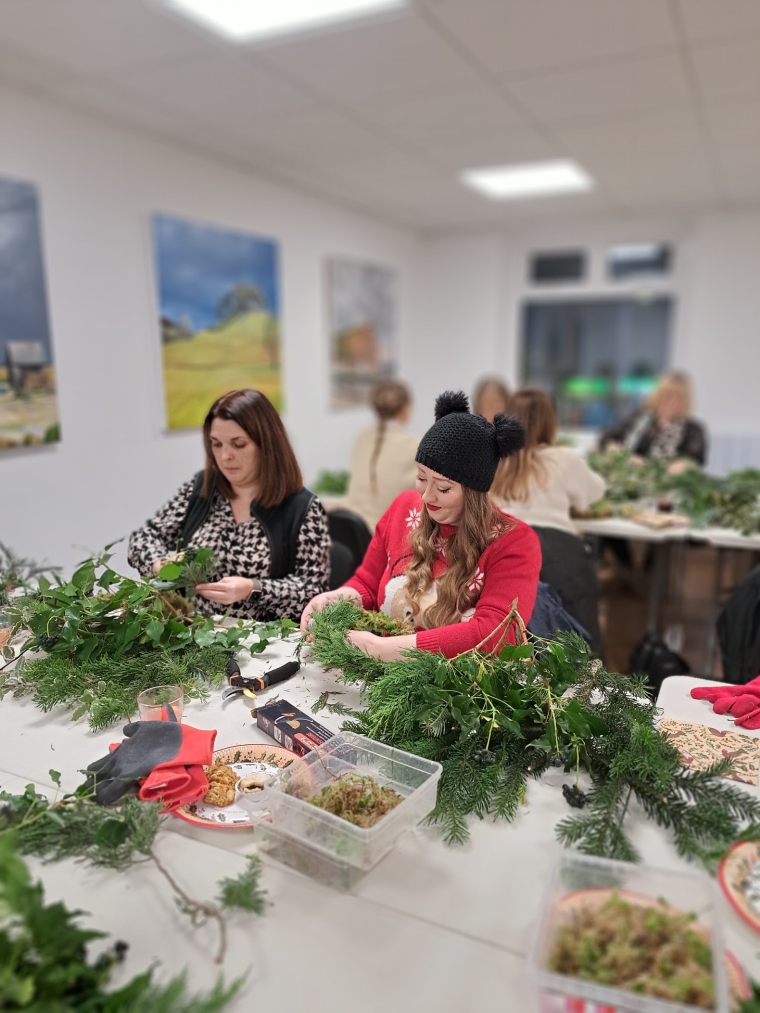 2 ladies sitting at a table constructing a Christmas wreath with lots of green foliage 