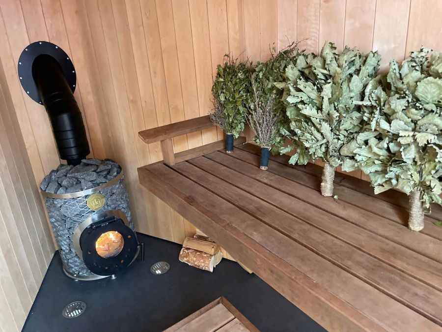 Wooden interior of a sauna with firebox, bench and bunches of leaves.