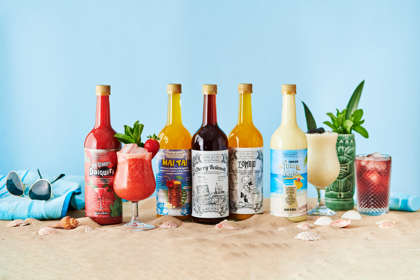 A selection of brightly coloured bottled cocktails and glasses with cocktails on sand with a towel and sunglasses to promote the summer cocktail range.