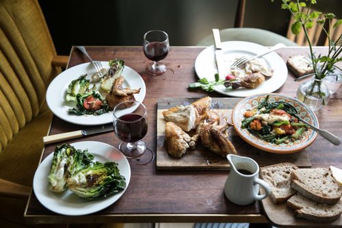 A dark wooden table with plates of food - roast chicken, vegetables and bread - and glasses of red wine.