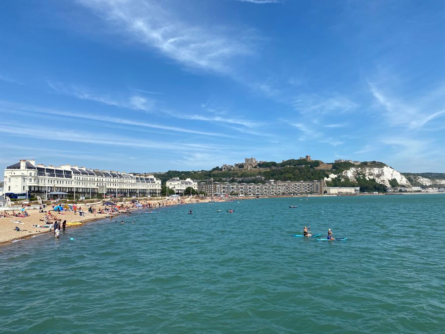 Dover Harbour beach with swimmers and paddle boarding with Dover Castle and the White Cliffs in the distance.
