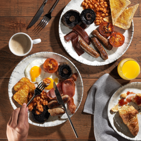 Two plates of cooked breakfasts showing sausages, bacon, tomatoes, eggs, baked beans and toast, on a wooden table. 