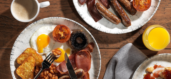 Two plates of cooked breakfasts showing sausages, bacon, tomatoes, eggs, baked beans and toast, on a wooden table. 