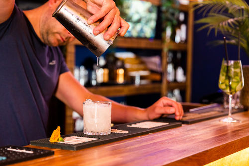 A man pouring a cocktail from a silver cocktail shaker into a glass on a wooden bar, bottles and glasses in the background.